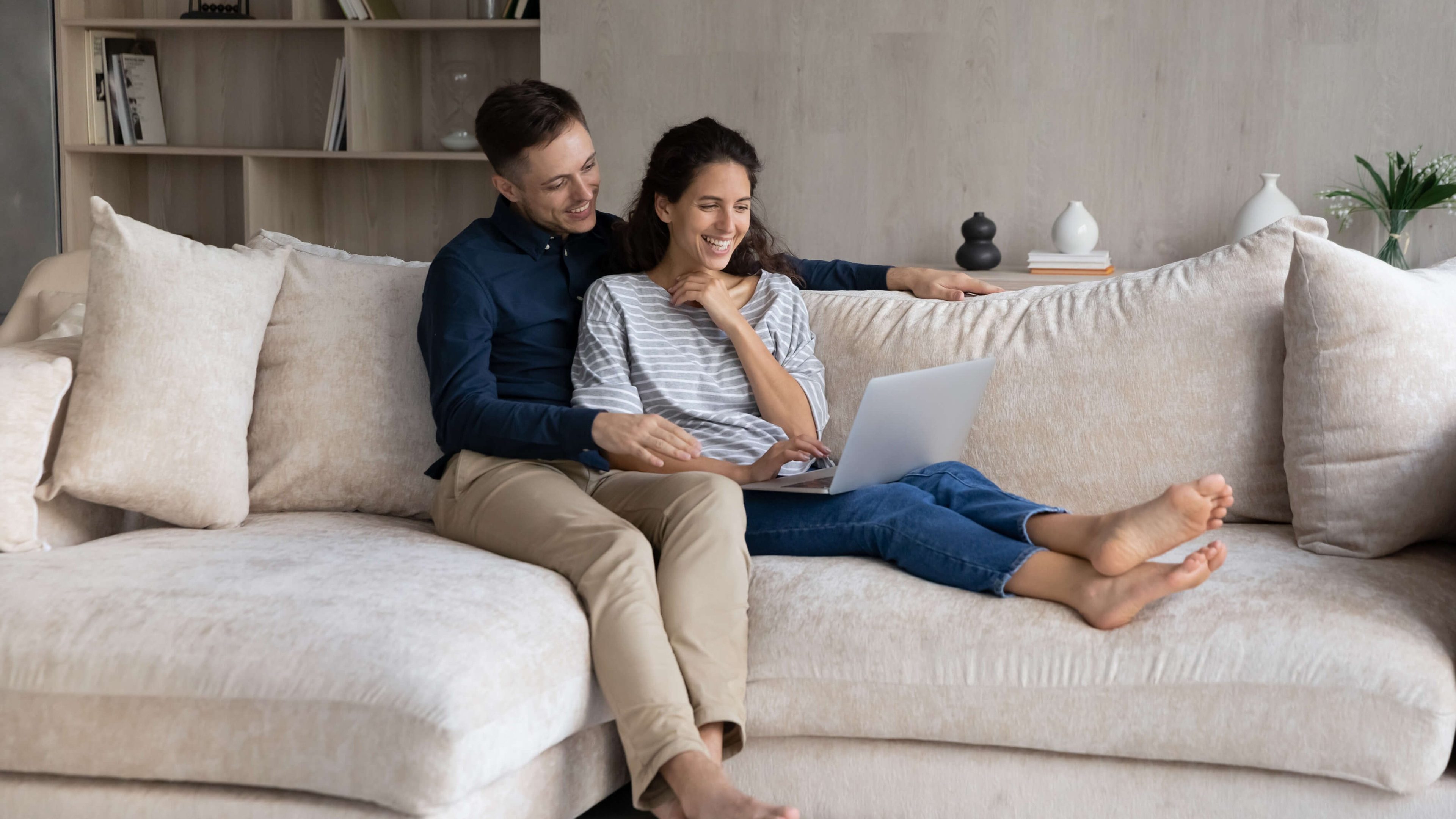 Happy sweet young couple watching movie on laptop, making video call, using online application, shopping online, looking at screen, laughing, sharing computer, relaxing on couch at home