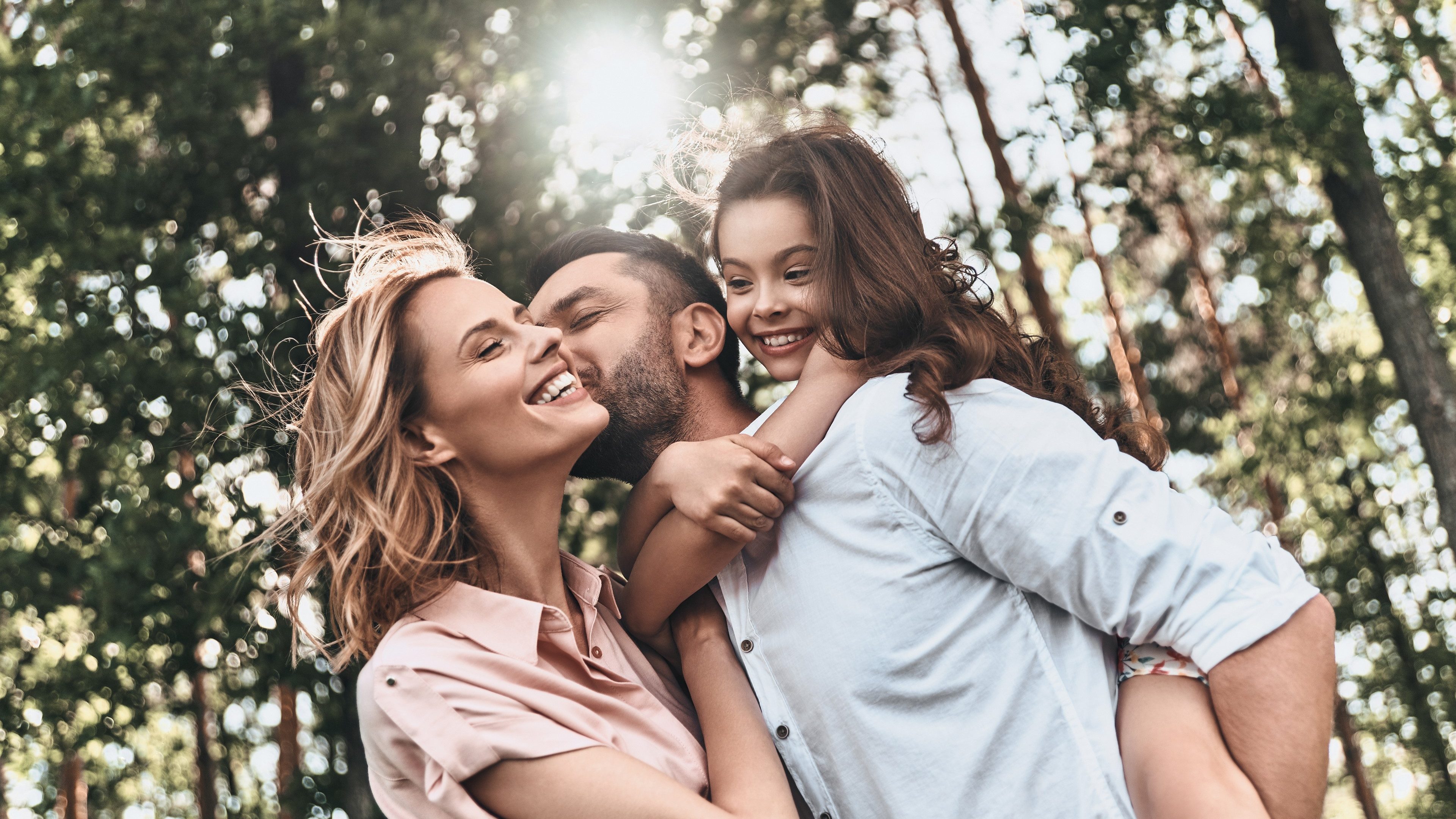 Enjoying time together. Young loving father carrying his smiling daughter and kissing his wife while spending free time outdoors