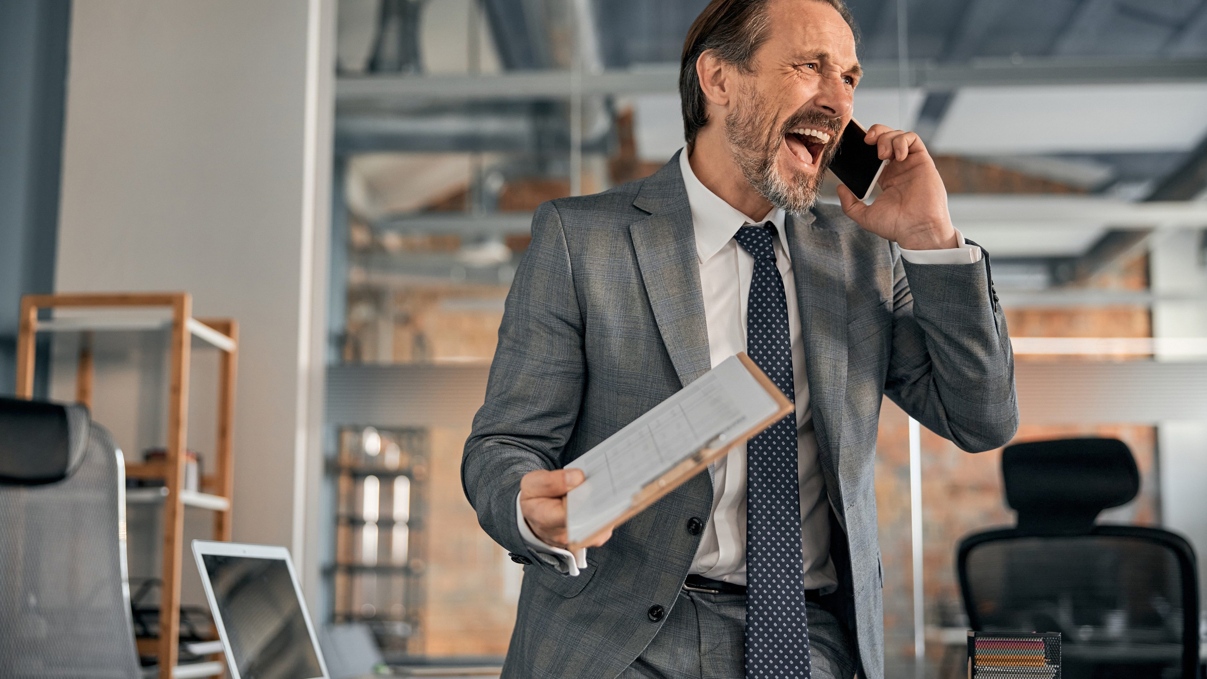 Happy bearded man having phone conversation and screaming with joy while sitting on office table