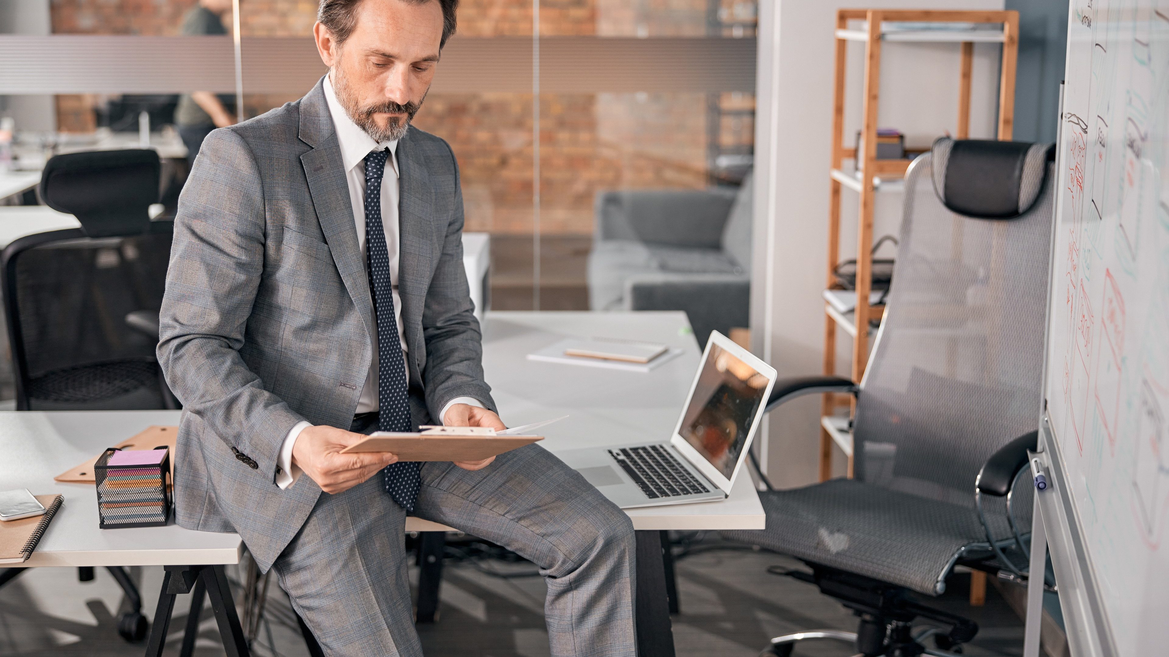 Handsome businessman in stylish suit sitting on office table and holding clipboard with papers