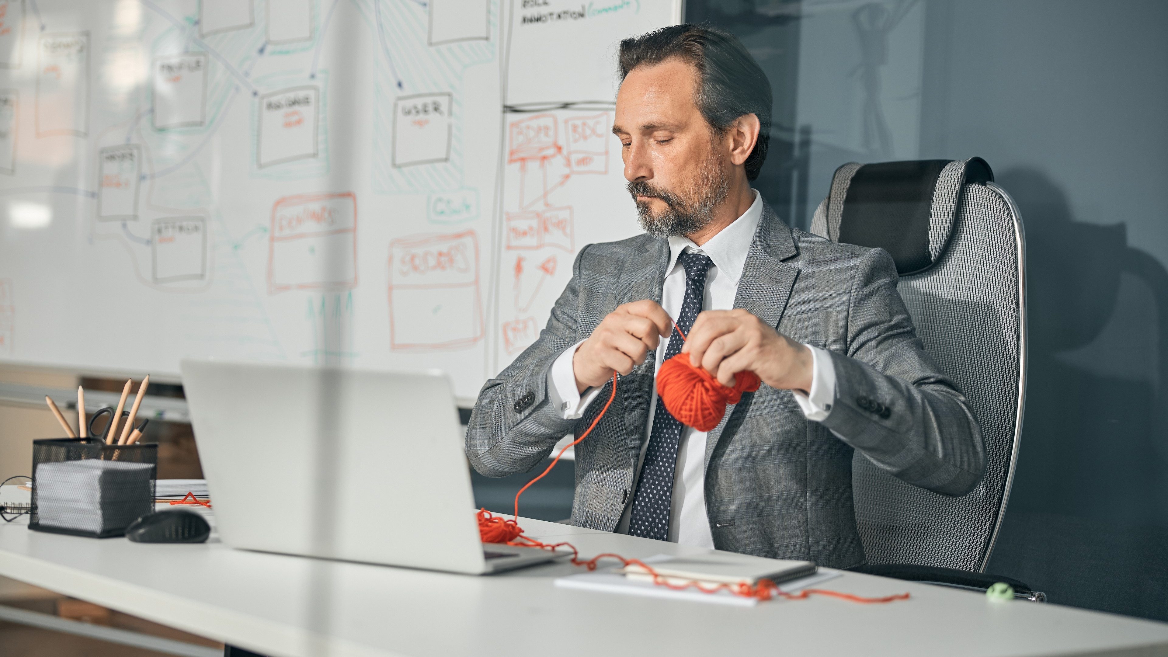 Concentrating office manager near laptop at office and holding ball of orange yarn in the hand