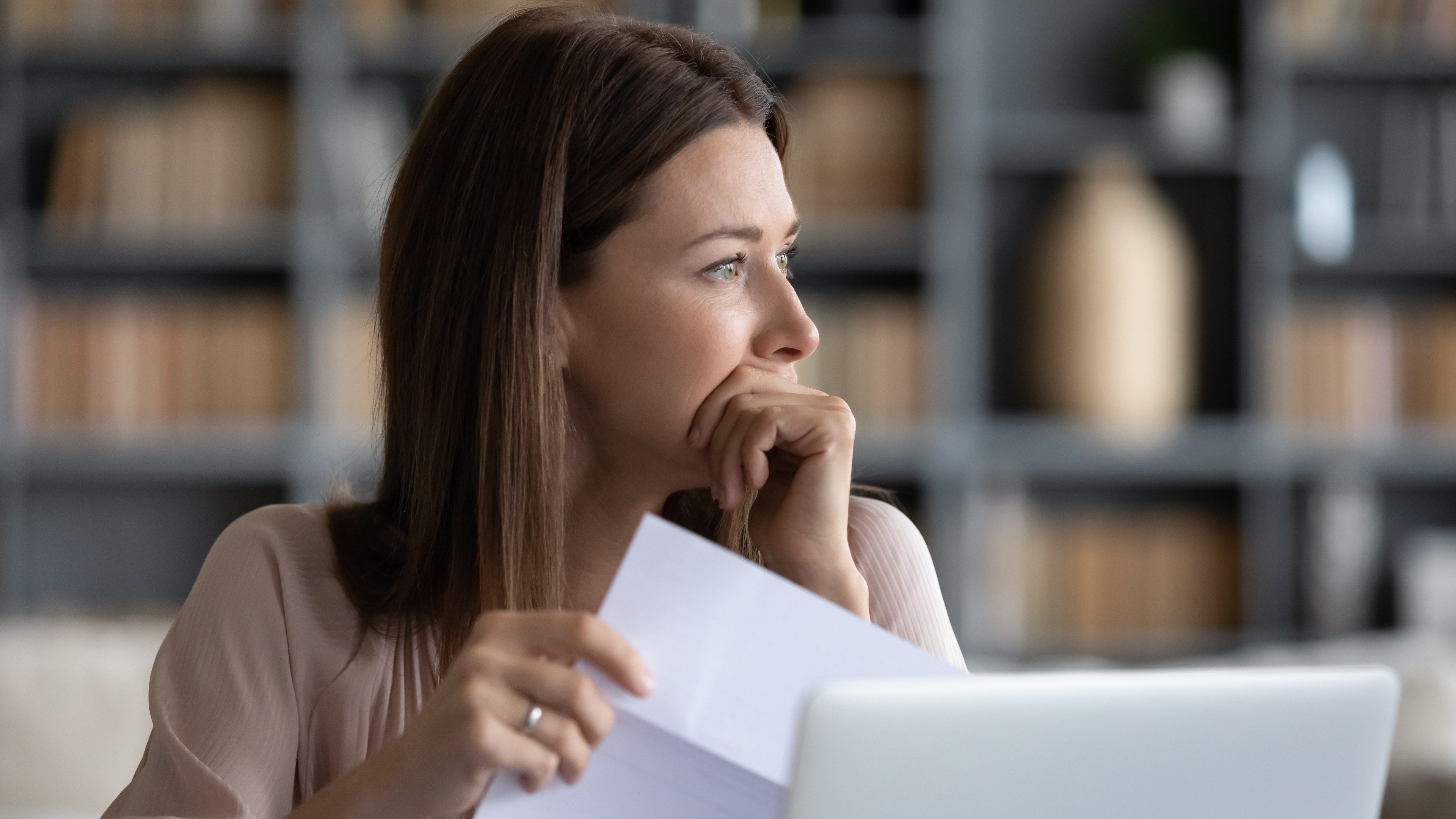 Head shot stressed young woman holding paper document, bank debt notification, thinking of financial troubles, looking away. Lost in negative thoughts depressed woman worrying about bad news notice.