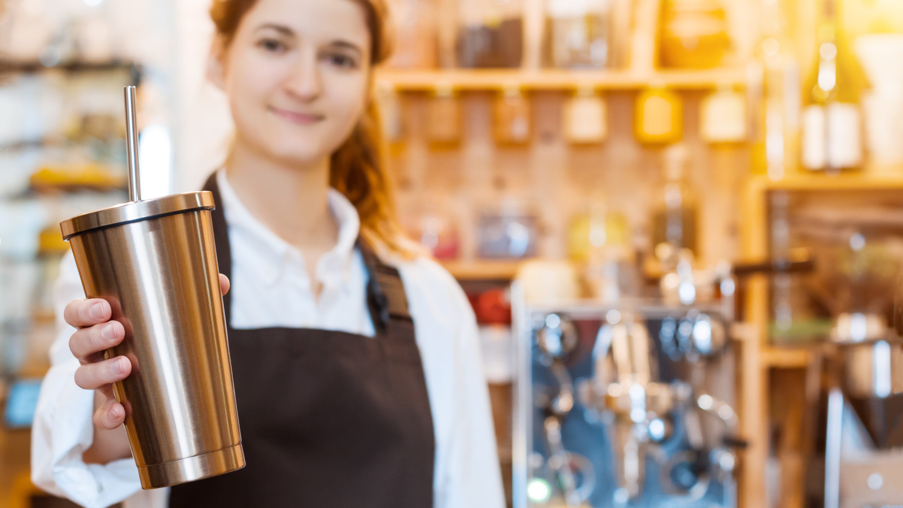 Closeup female hands are holding stainless metal reusable tumbler cup mug with drinking straw. Barista woman  prepared, brewed coffee using professional machine in cafe, restaurant. Take away, to go.