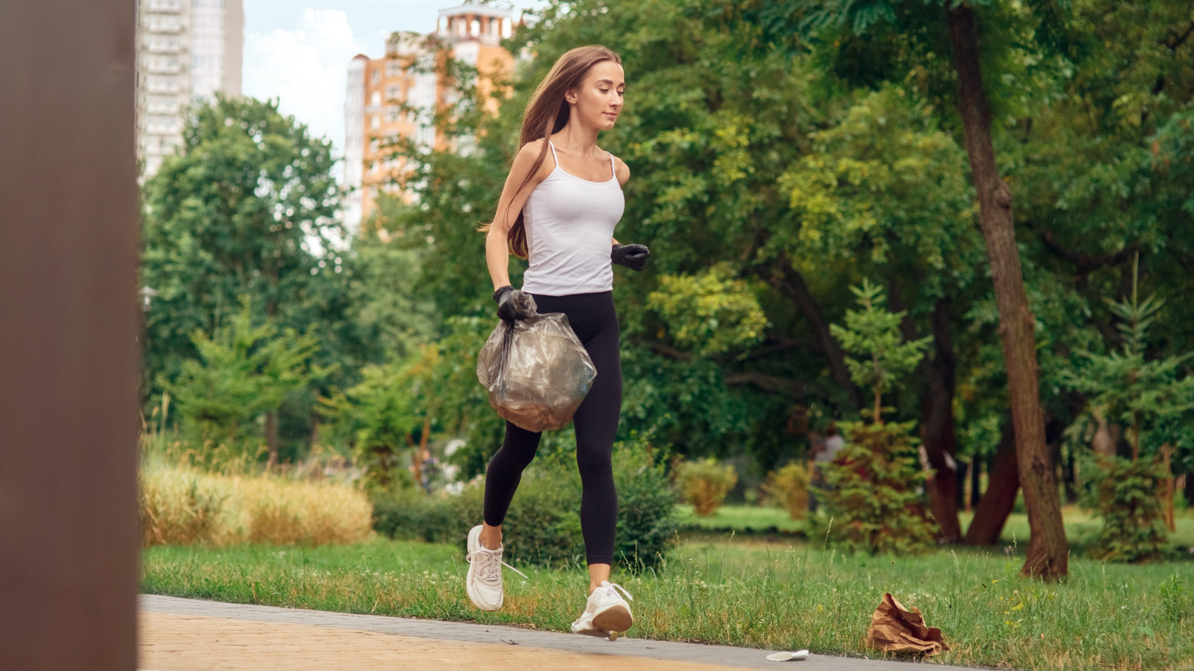 Young woman volunteer outdoors running wearing gloves in the park helping nature plogging holding plastic bag picking up litter smiling pensive