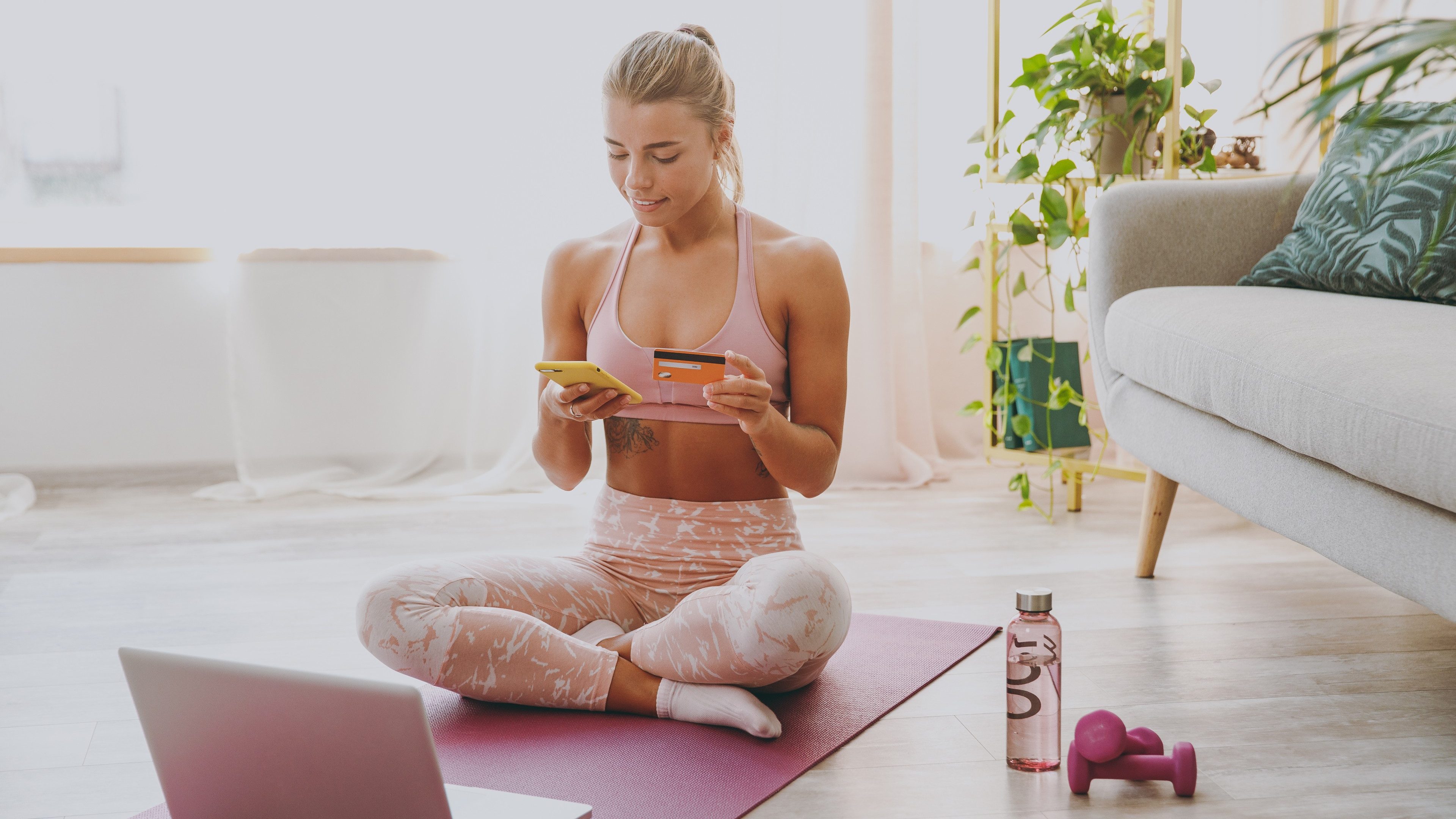 Pretty young strong sporty fitness woman wearing pink tracksuit doing yoga exercises using laptop mobile phone hold credit bank card showing thumb up sitting stretching on mat floor at home indoor
