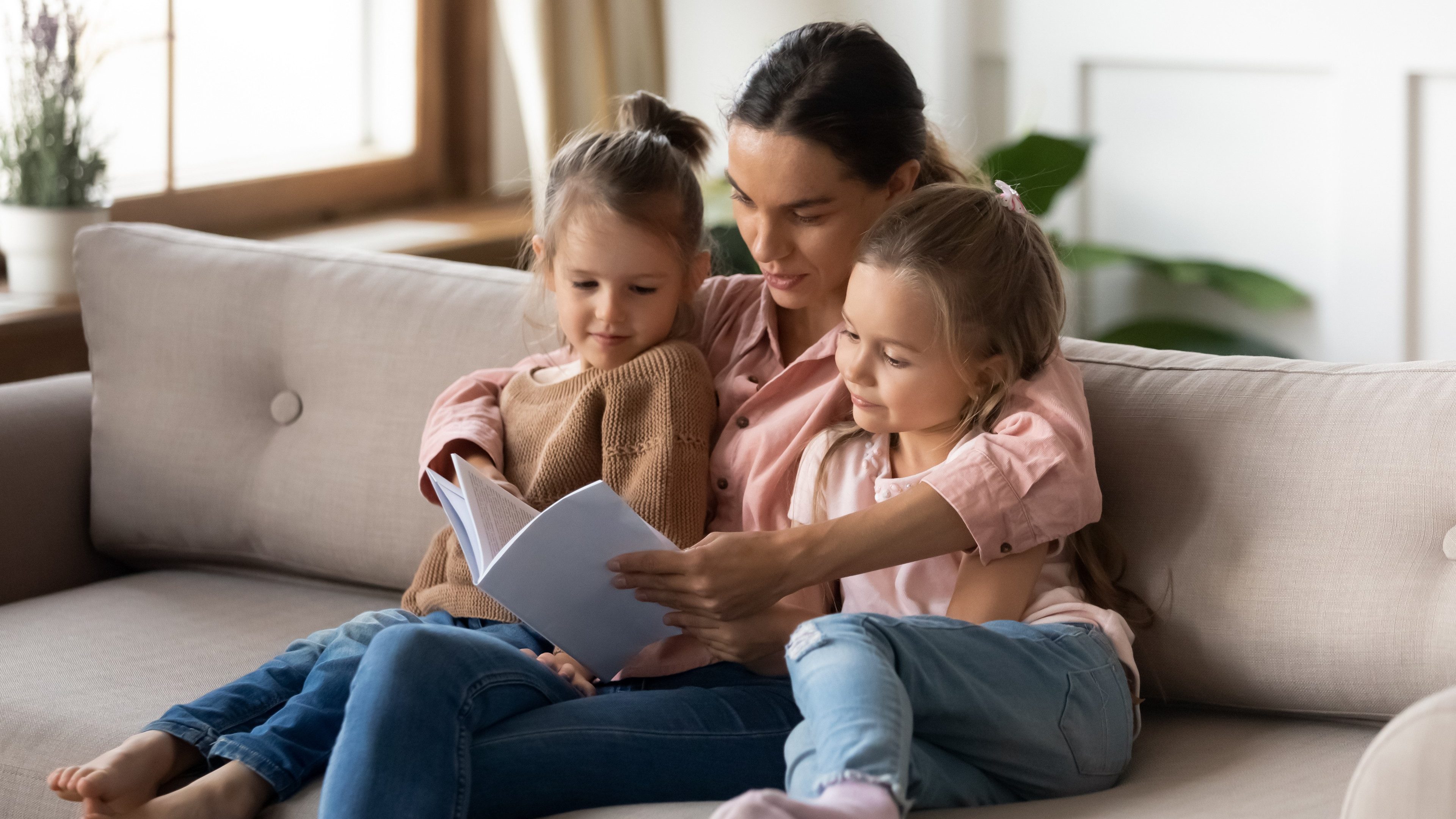 Two cute daughters enjoy interesting fairy tale story read by mommy, sitting together on couch in living room. Hobby and leisure funny time, development and education of children by parent at home