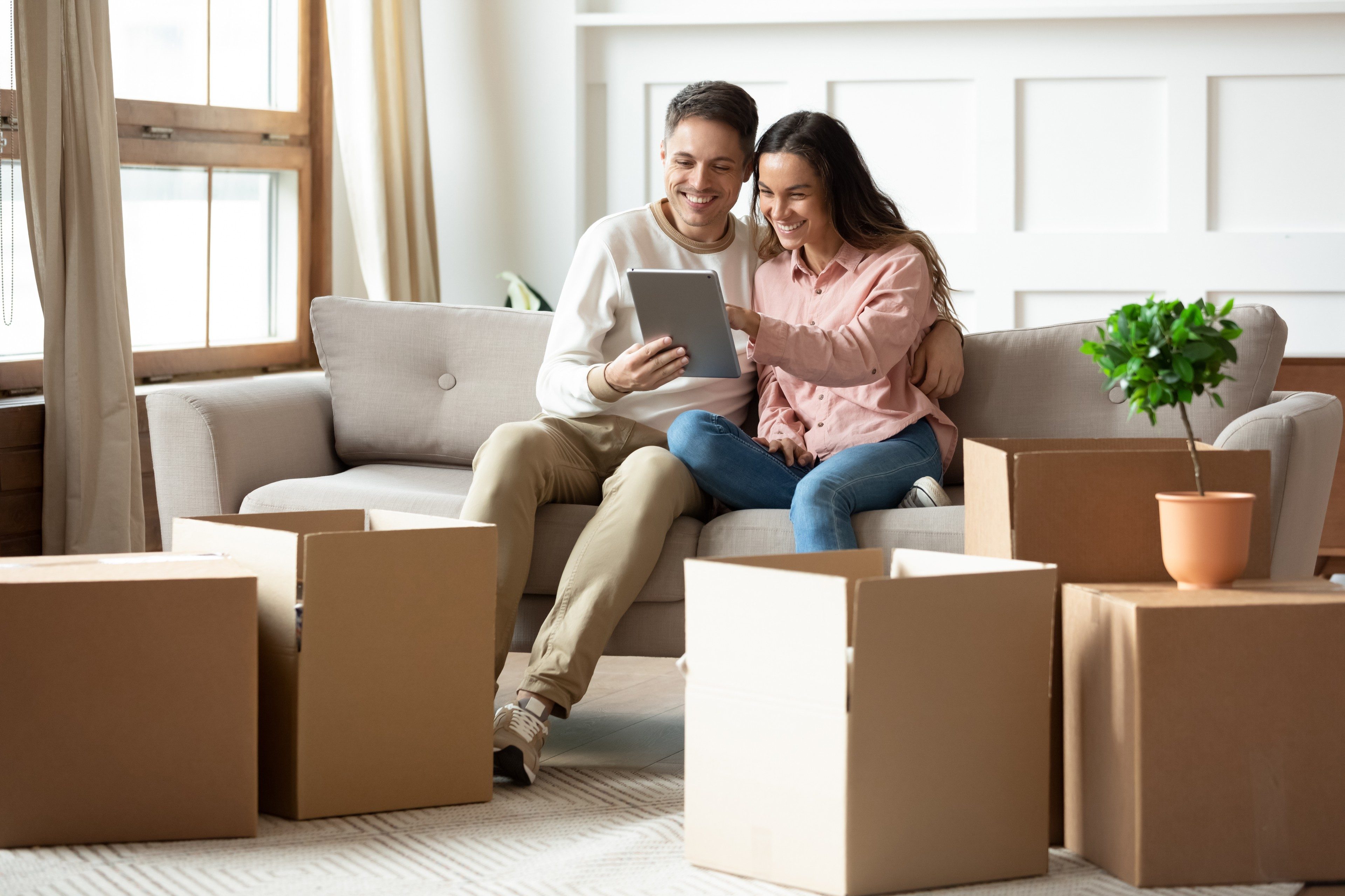 Happy couple sitting on sofa surrounded by heap of cardboard boxes, using tablet choose goods and furniture for new home, resting at moving day at first dwelling. E-commerce retail services concept