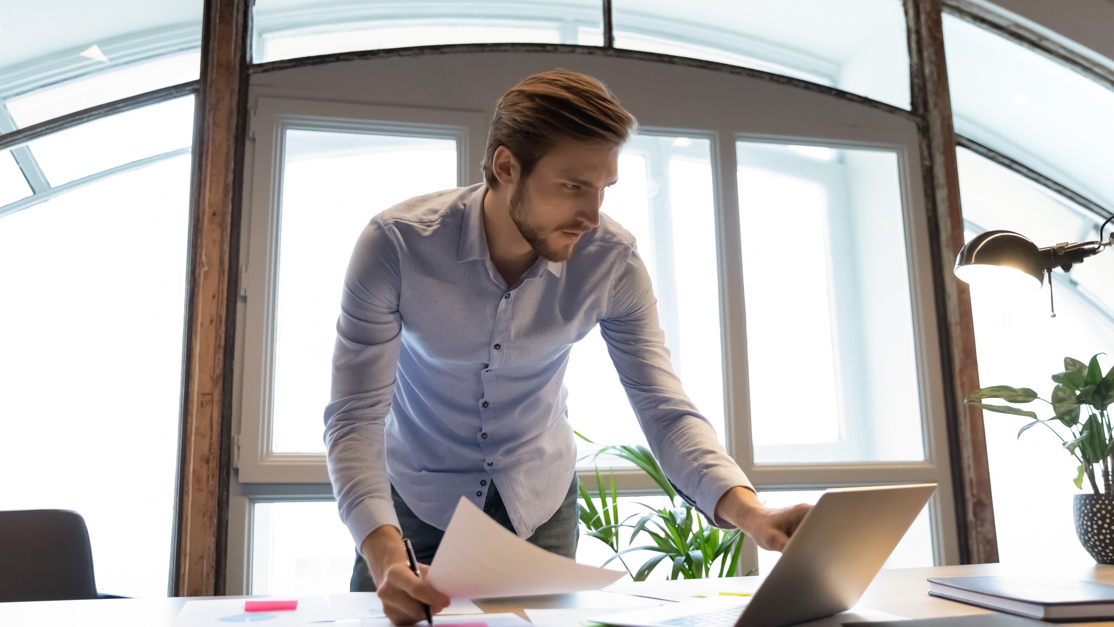 Concentrated young male project manager software developer standing by desk at workplace in office involved in paperwork, checking comparing data in hardcopy and on laptop screen, making notes changes