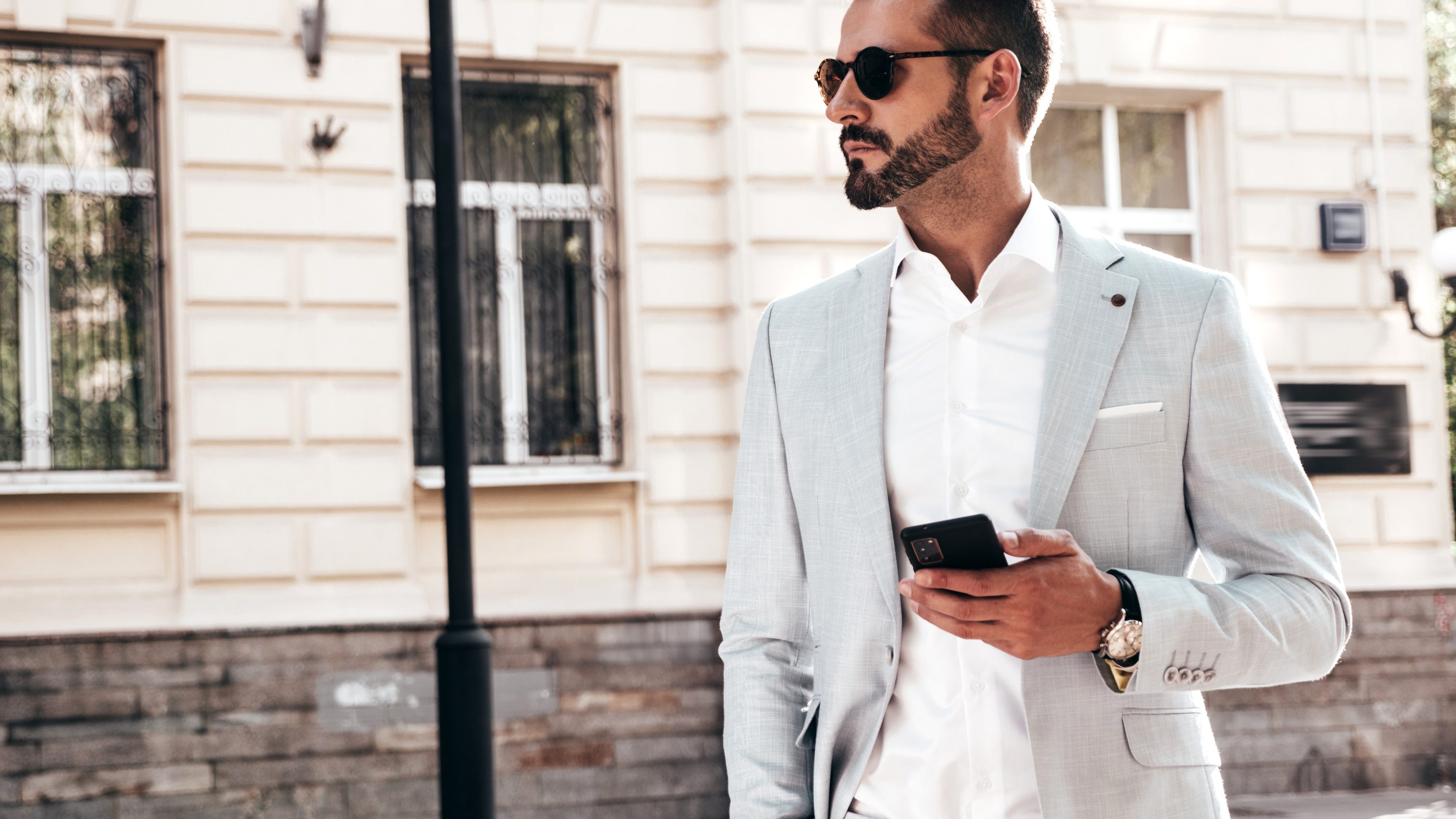 Portrait of handsome confident stylish hipster lambersexual model.Sexy modern man dressed in elegant suit. Fashion male posing in the street near fence at sunset. In sunglasses.Using smartphone apps
