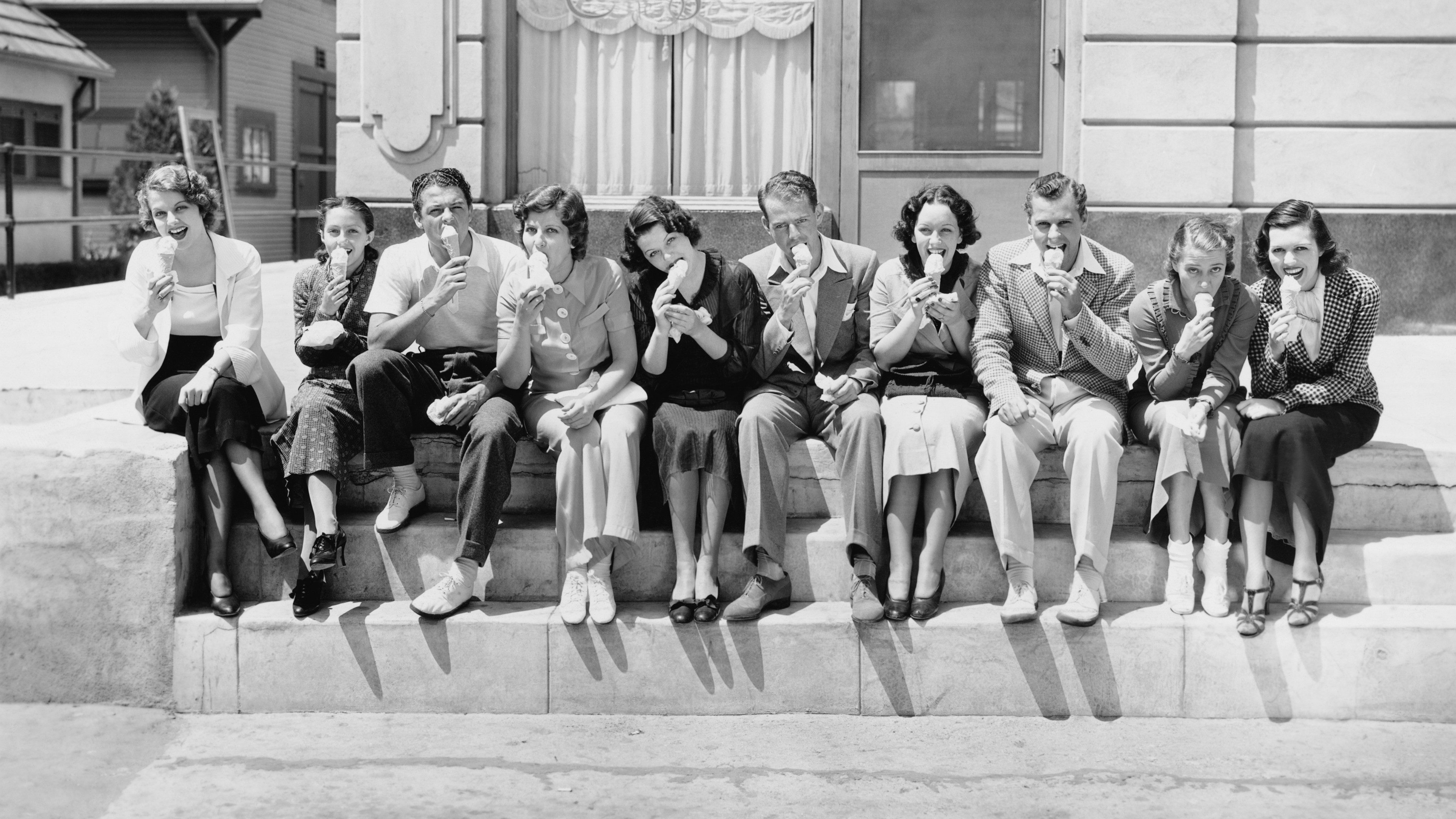 Historická fotografia z cca 60. rokov: Partia mladých ľudí sedí na schodoch pred budovou a líže zmrzlinu.