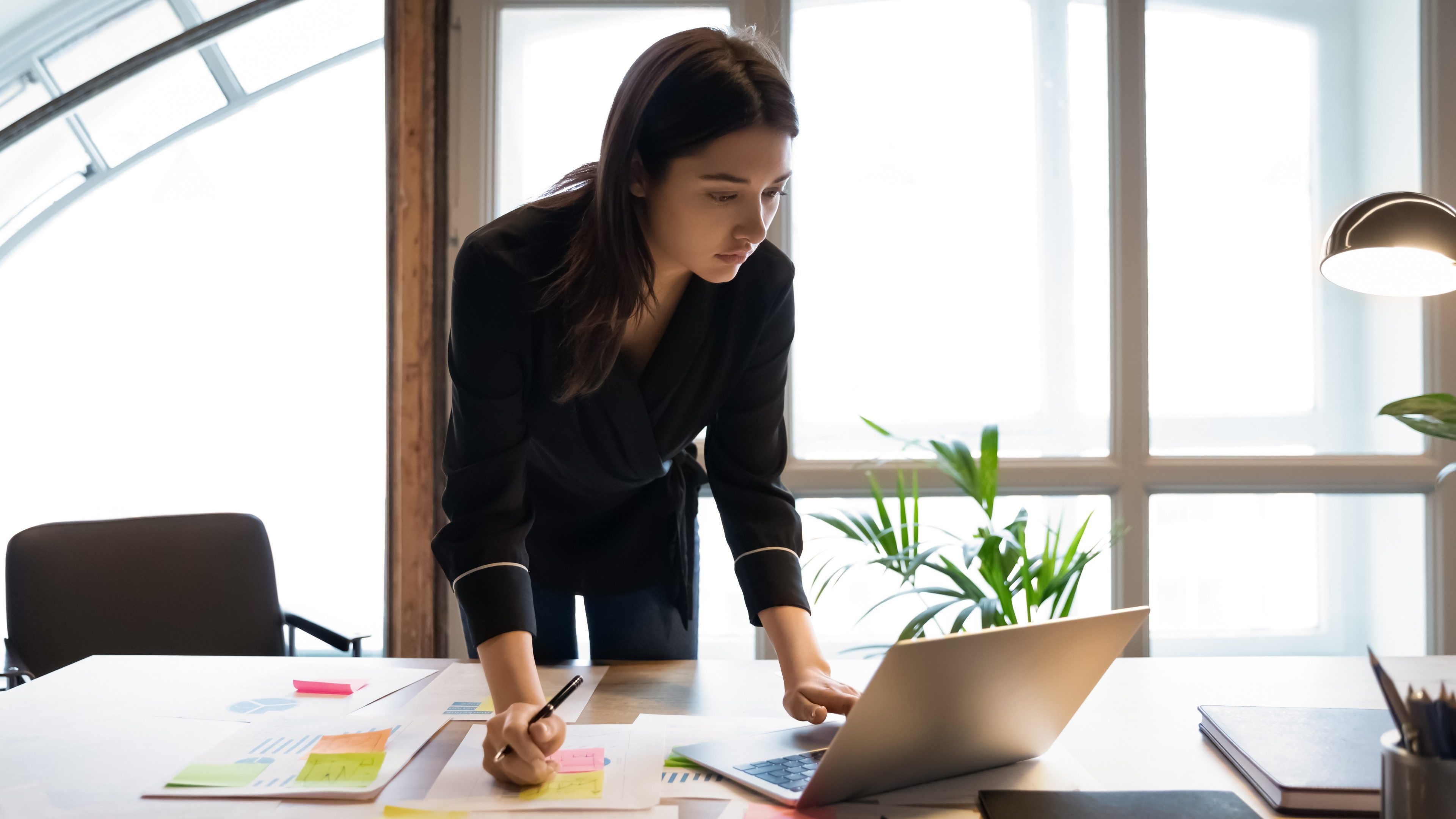 Confident millennial indian female business analyst financial advisor preparing statistic report studying documents on work desk, browsing information online using pc, writing out notes on paper sheet