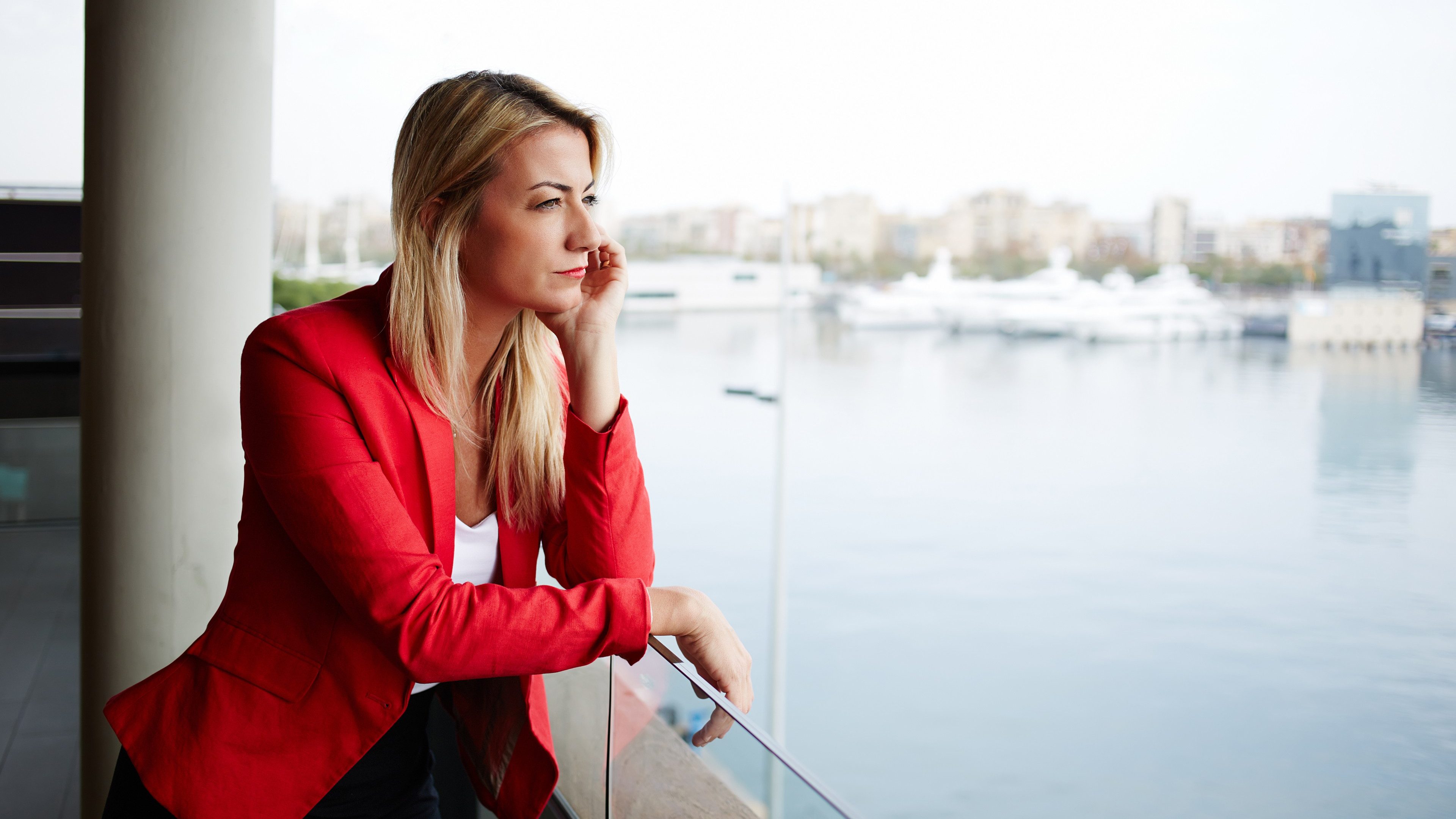 Portrait of pensive businesswoman looking out of an office balcony with beautiful seaport view on background, unhappy successful woman standing in exterior,blonde woman looking out of her window sadly