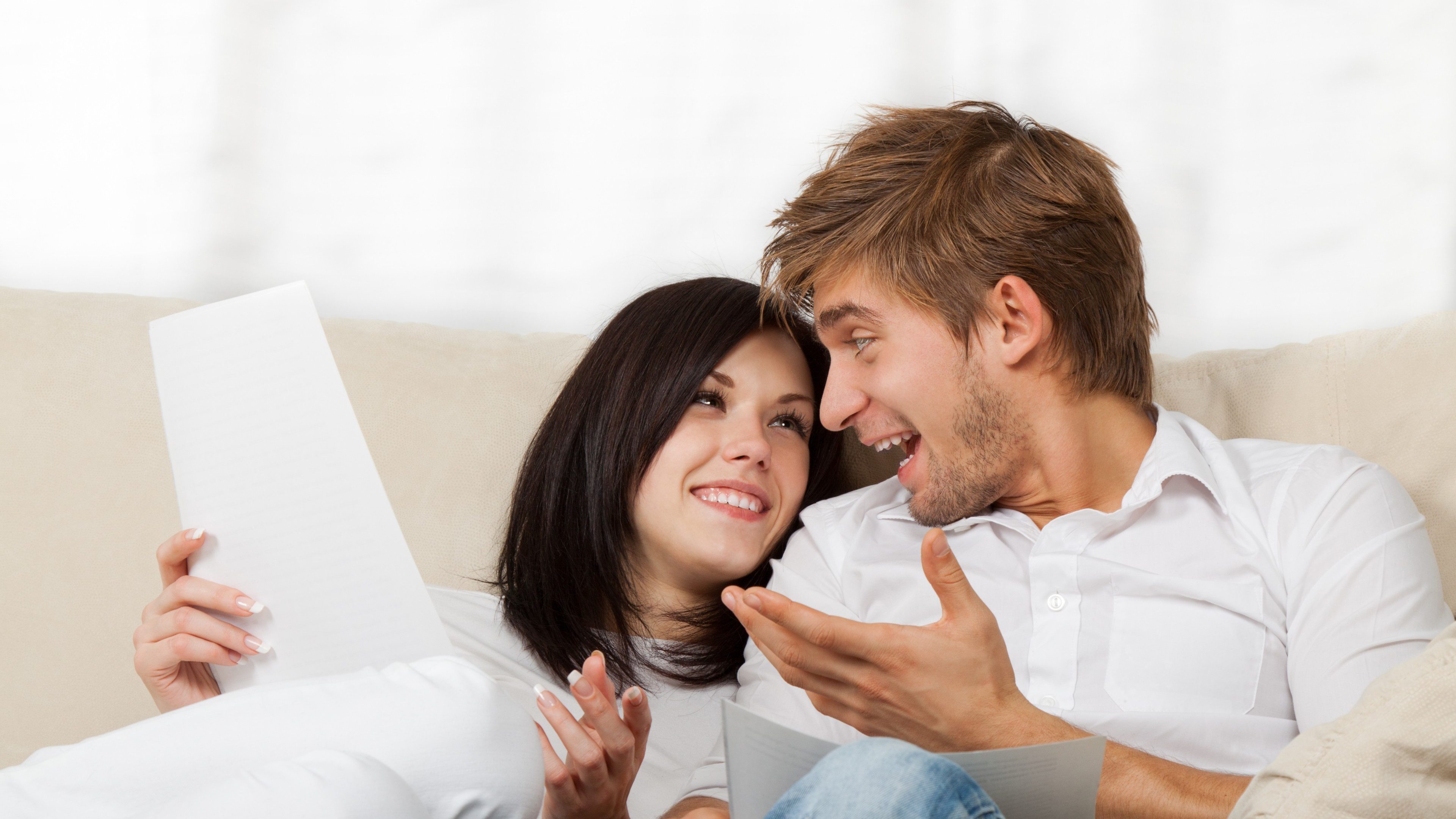 Happy couple reading a letter in their living room, young smile man and woman sitting on couch, sofa looking to each other talking