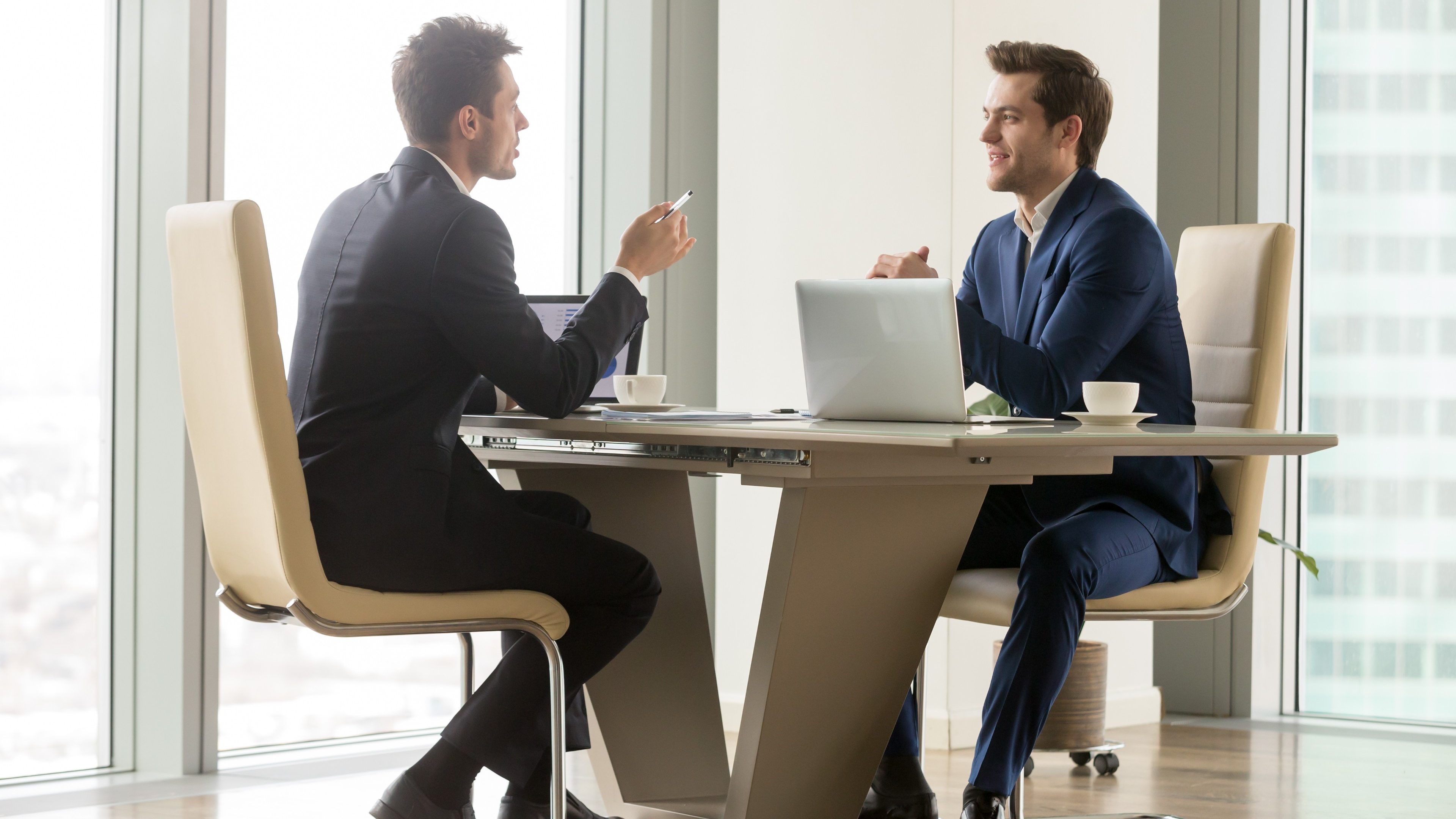 Two handsome businessmen sitting in comfortable chairs at desk with laptops in meeting room. CEO making important negotiation about companies partnership or corporate merger. Financiers planning deal 