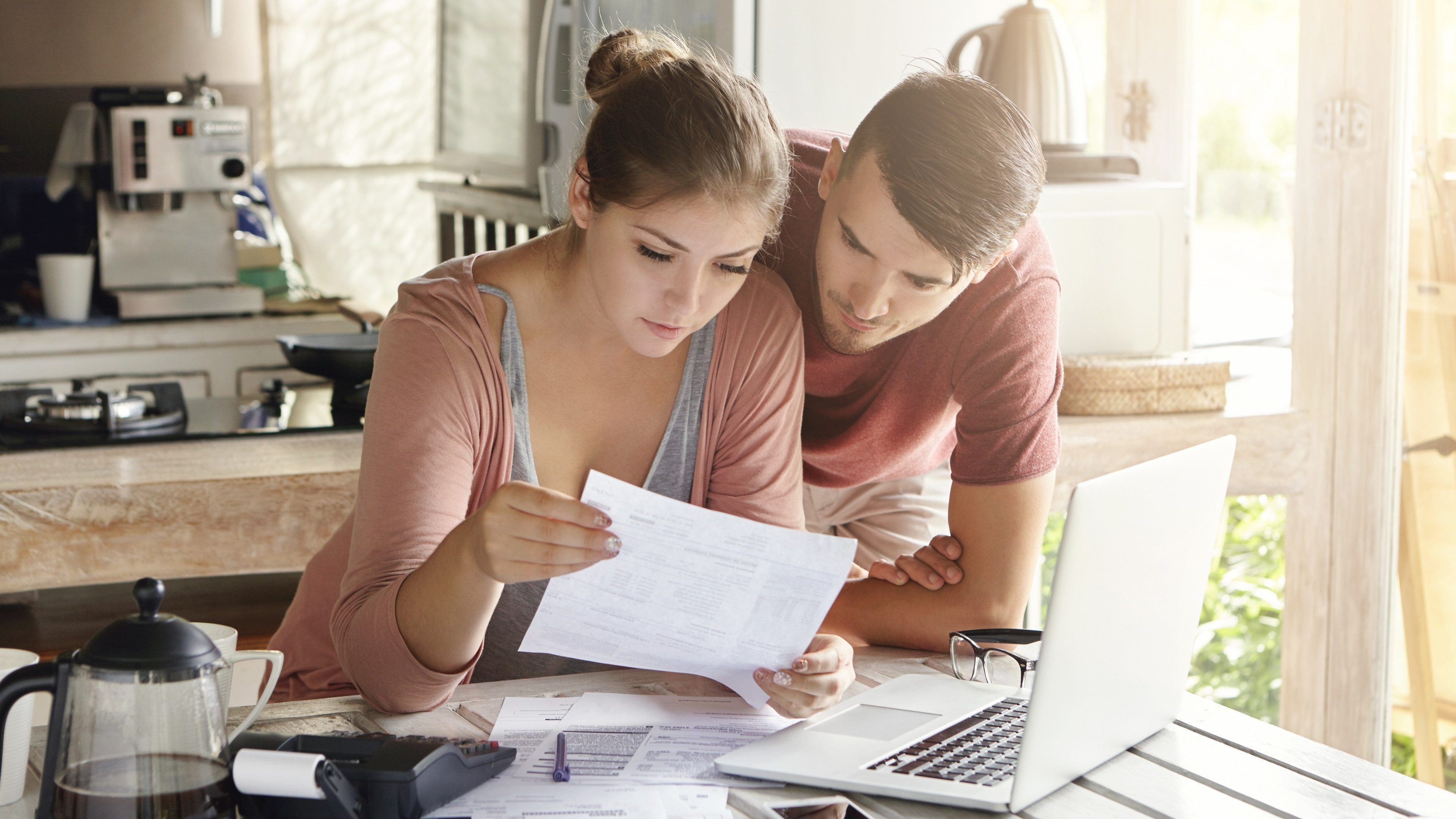 Young couple managing finances, reviewing their bank accounts using laptop computer and calculator at modern kitchen. Woman and man doing paperwork together, paying taxes online on notebook pc