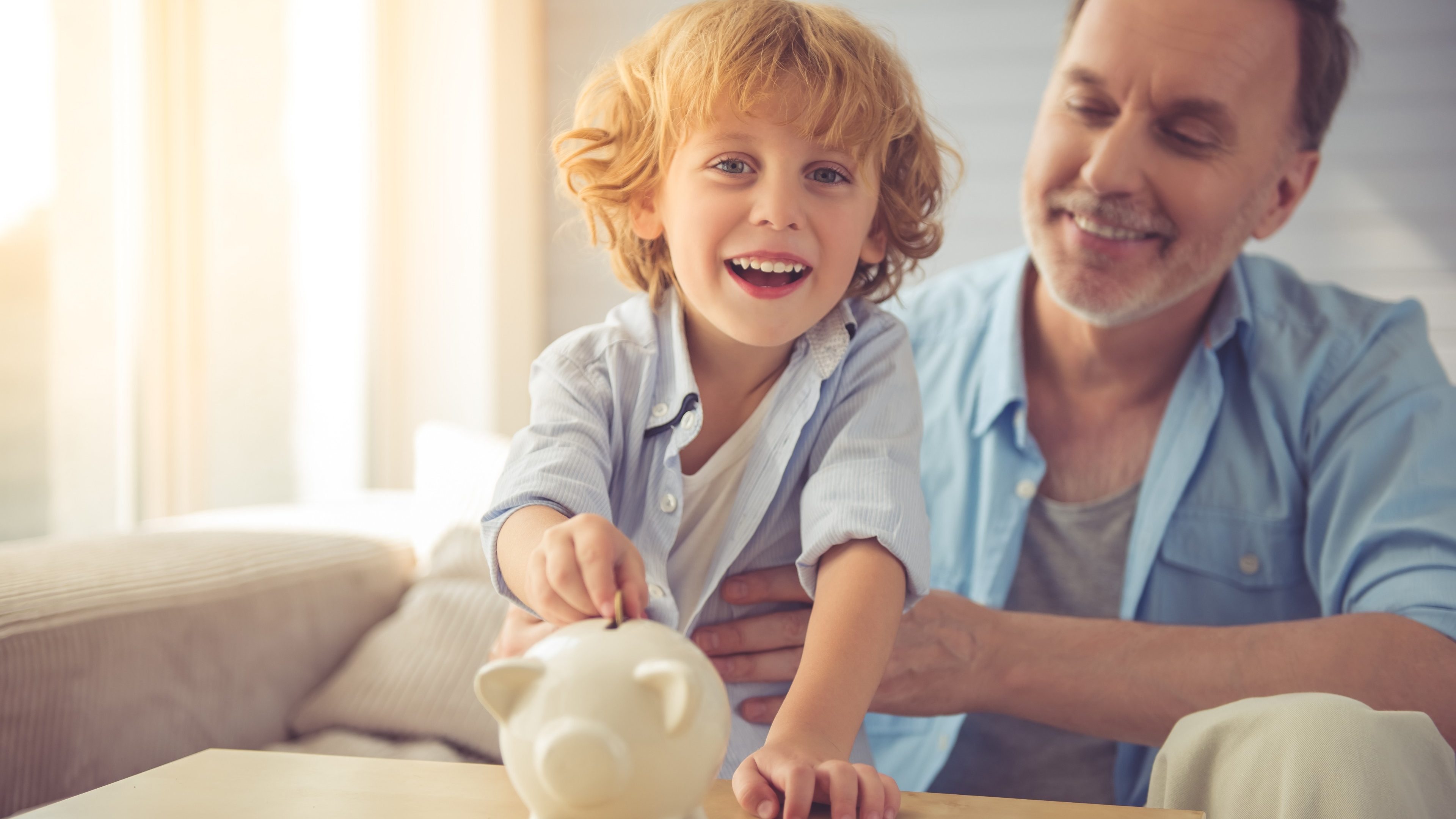 Cute little boy is putting coin into the piggy bank, looking at camera and smiling while spending time with his grandpa at home