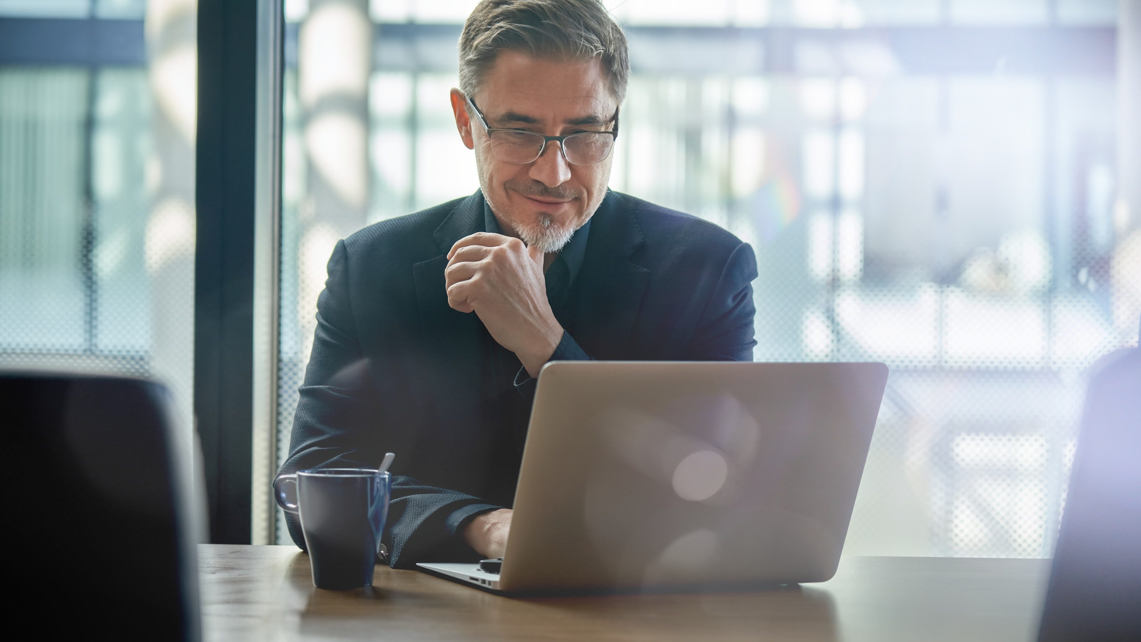 Business portrait - businessman sitting in in office working with laptop computer. Mature age, middle age, mid adult man in 50s with happy confident smile. Copy space.