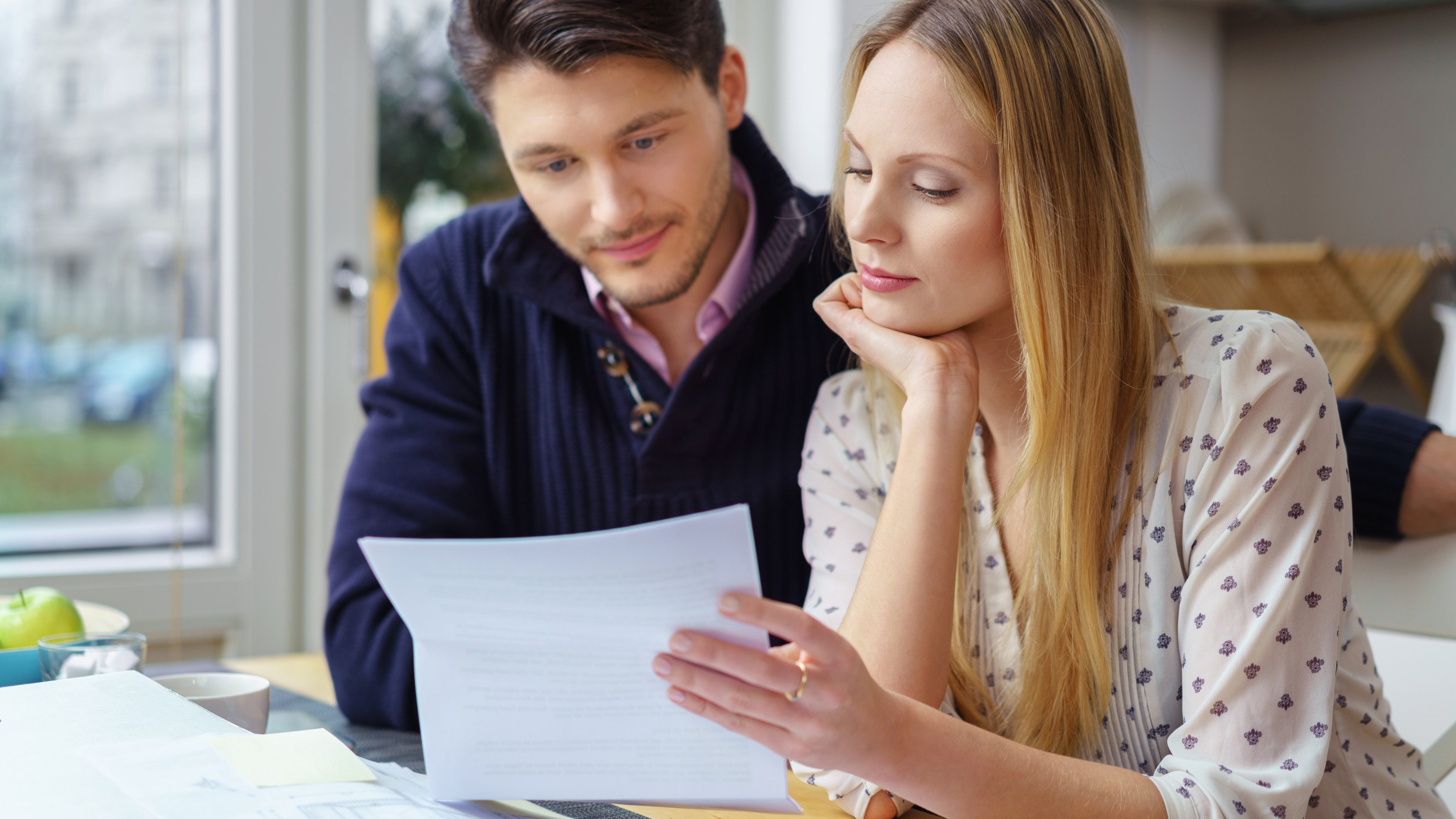 Handsome young man with mustache and beautiful woman with long hair at table looking at documents in kitchen next to window