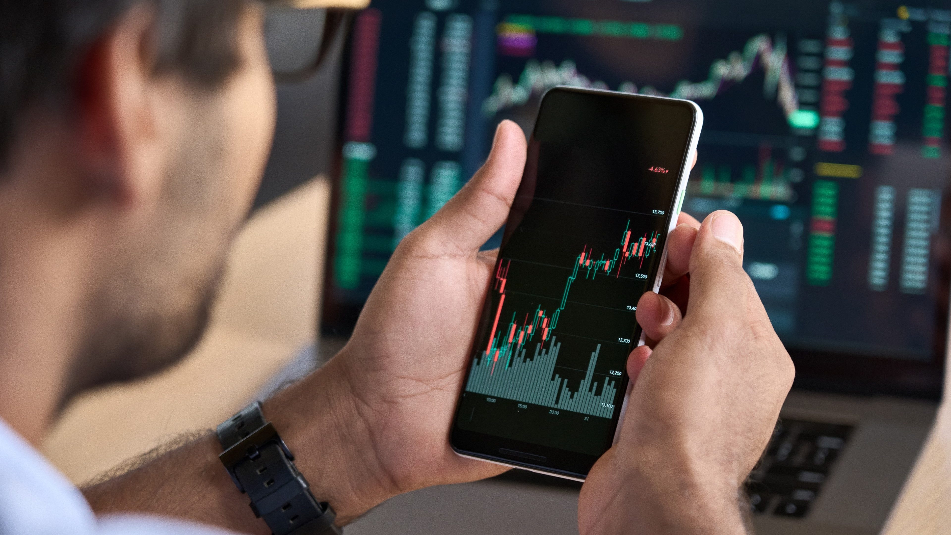 Male trader investor broker crypto analyst holding smartphone in hand analyzing stock market trading charts indexes data checking price using mobile stockmarket exchange app, over shoulder view.