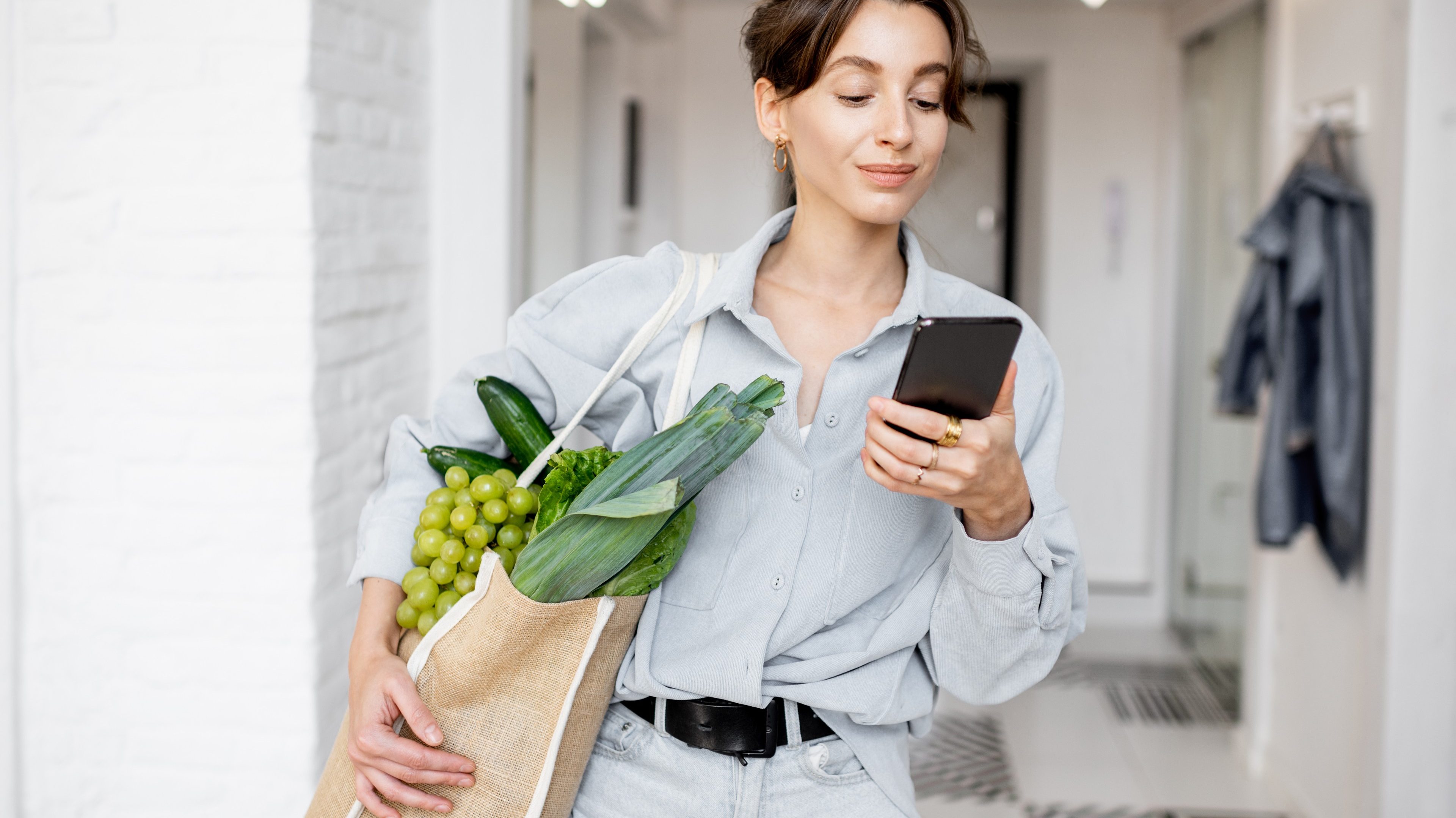 Portrait of a young and cheerful woman standing with mobile phone and shopping bag full of fresh food at home. Concept of buying products online